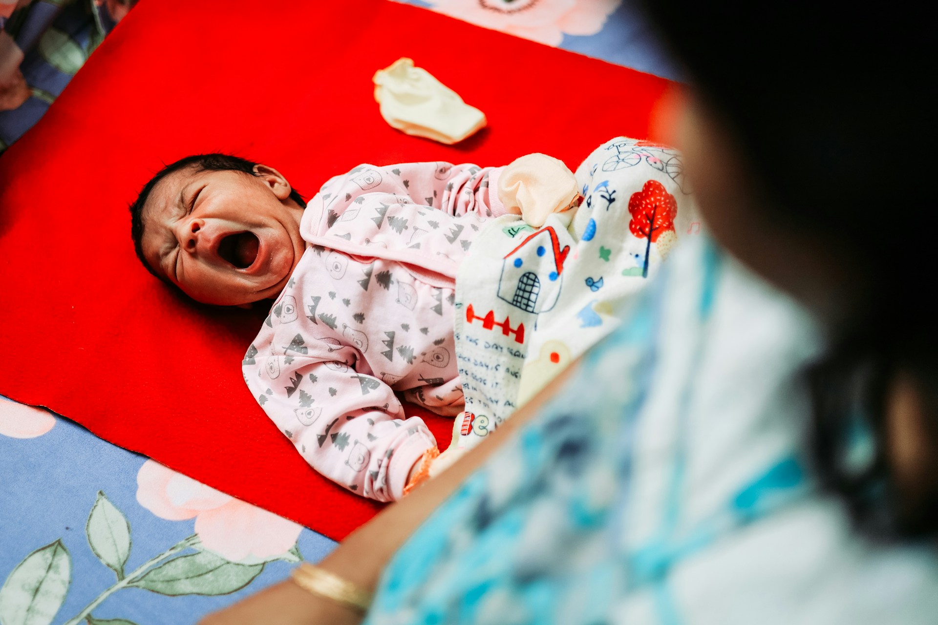A newborn yawning, wearing a pink onesie with tree patterns, as his mother looks on from the side