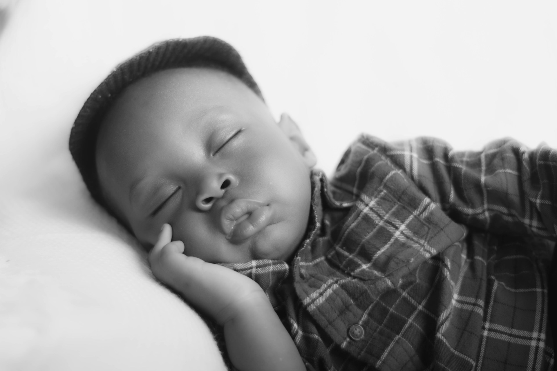 Black and white portrait of a sleeping newborn wearing a knit cap and plaid shirt